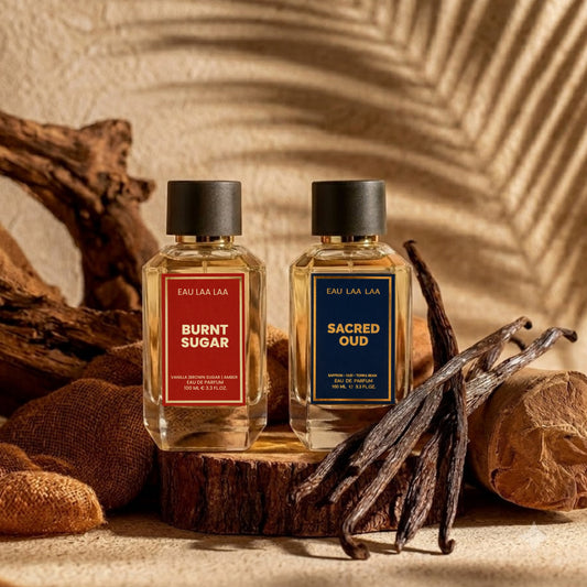 Two perfume bottles labeled 'Burnt Sugar' and 'Sacred Oud' on a wooden surface with vanilla beans.