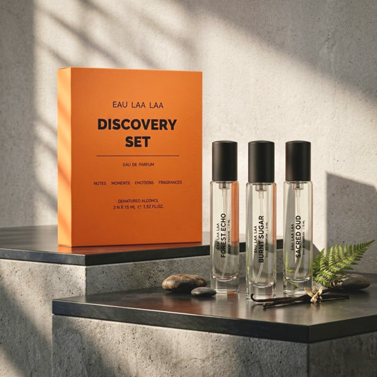 Eau Laa Laa Discovery Set perfume gift set featuring an orange box and three clear glass sample vials with black caps, styled on a dark stone surface against a textured gray wall with dramatic shadows.