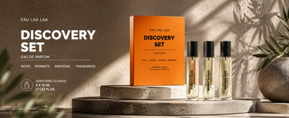 Eau Laa Laa Discovery Set perfume, featuring an orange box and three clear glass bottles with black caps, displayed on stone pedestals with warm, shadowed lighting.